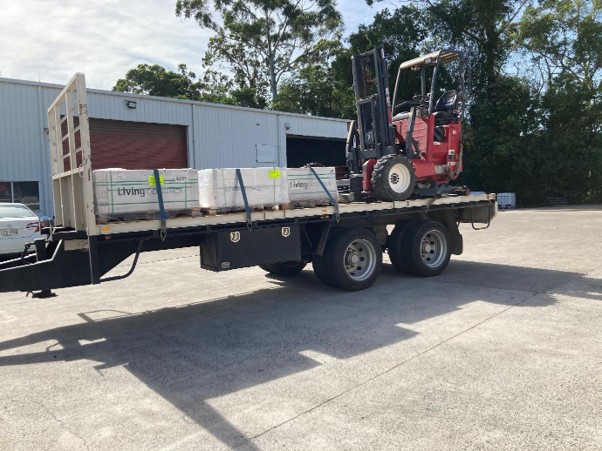 Flatbed East Coast Distribution freight truck with a truck-mounted forklift unloading palletised goods (Living Ceramics) at an industrial warehouse yard, demonstrating mobile forklift hire for on-site delivery where no loading dock is available.